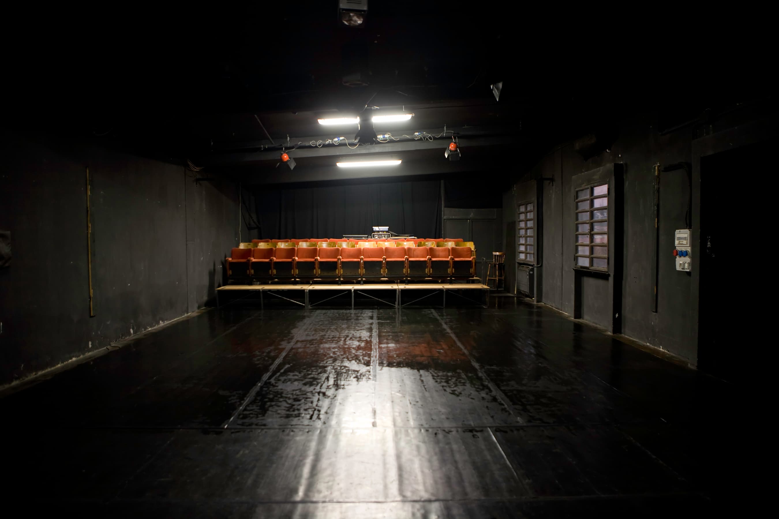 Dimly lit theater with a row of empty orange seats facing a dark, glossy stage floor. Overhead lights illuminate the seating area.